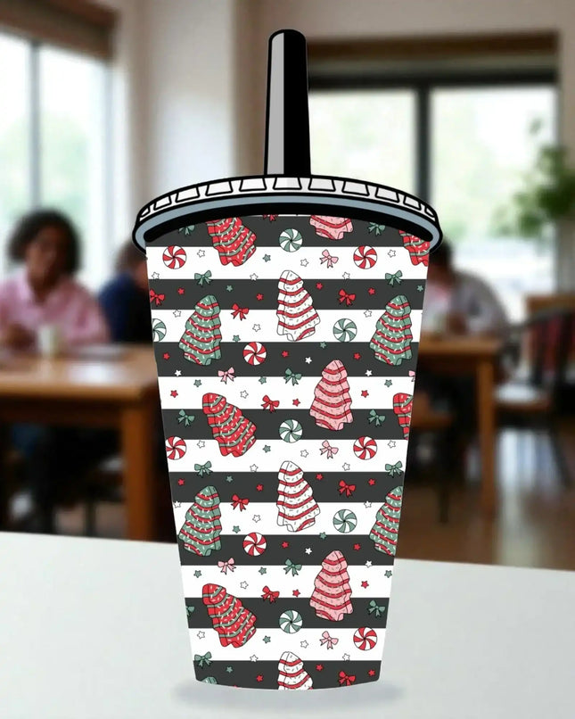 A colorful, patterned cup with Christmas-themed designs, including trees, candy canes, and other festive elements, is displayed on a table.